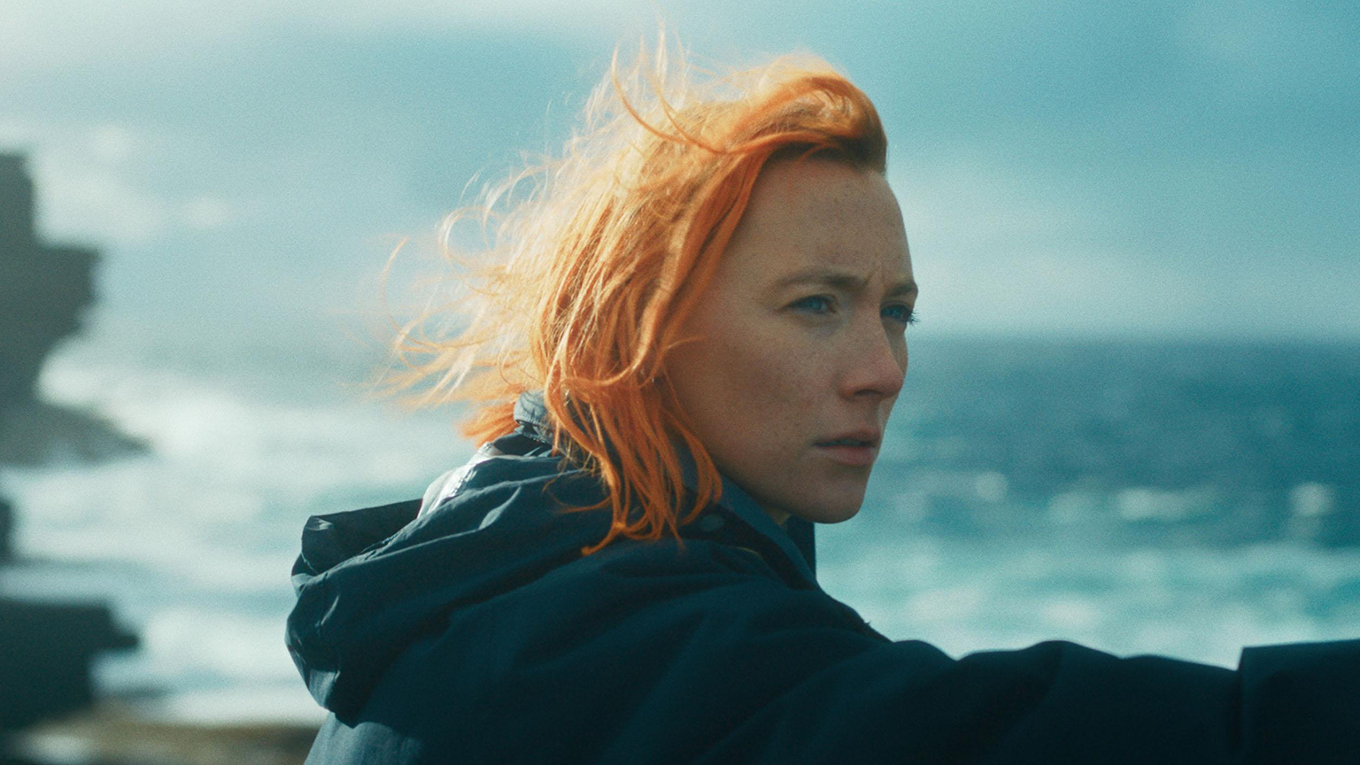 A woman with orange hair by the ocean looking in the distance.