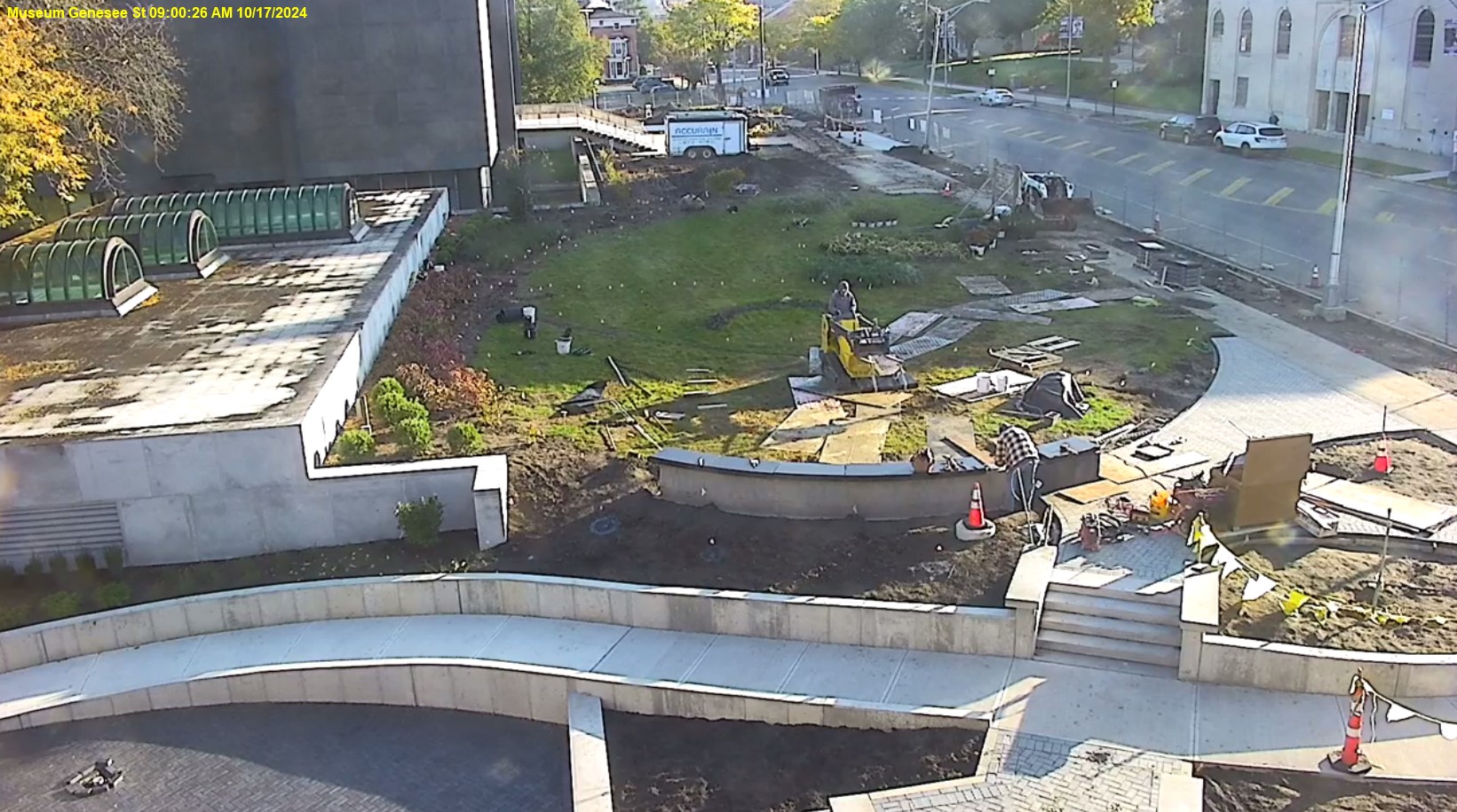 View of Genesee Street front lawn with the museum in the top left corner, the roof of the education wing middle of the left side, street on the right, and showing construction progress in the middle, the concrete sides of the ramp are in progress.