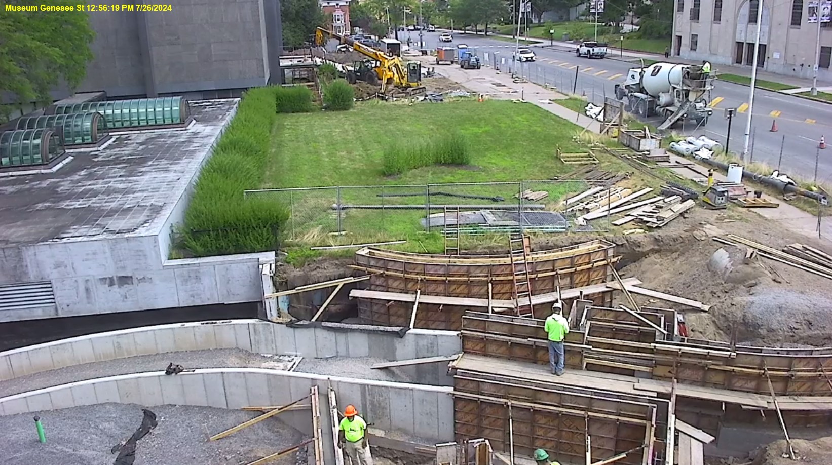 View of Genesee Street front lawn with the museum in the top left corner, the roof of the education wing middle of the left side, street on the right, and showing construction progress in the middle, the concrete sides of the ramp are in progress.
